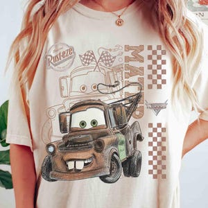 May include: Beige t-shirt featuring a cartoon illustration of Mater from the movie Cars. The design includes Mater's face, the word "Mater", checkered flags, and the "Rust-eze" logo. The shirt has a crew neck and short sleeves.