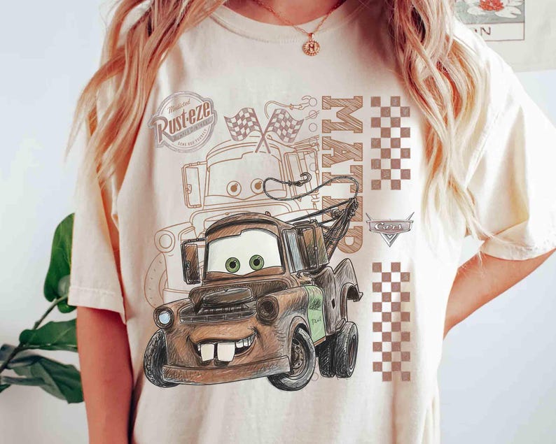 May include: Beige t-shirt featuring a cartoon illustration of Mater from the movie Cars. The design includes Mater's face, the word "Mater", checkered flags, and the "Rust-eze" logo. The shirt has a crew neck and short sleeves.