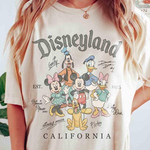 Puede incluir: Camiseta color crema con el logotipo de Disneyland y varios personajes de Disney, incluyendo Mickey Mouse, Minnie Mouse, Donald Duck y Goofy. La camiseta tambi&eacute;n incluye el texto "CALIFORNIA" y el a&ntilde;o "1955".