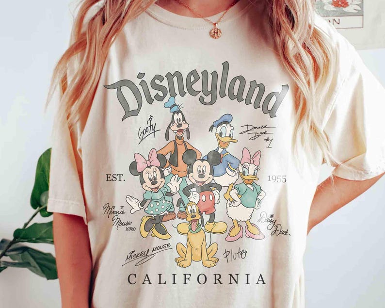 Puede incluir: Camiseta color crema con el logotipo de Disneyland y varios personajes de Disney, incluyendo Mickey Mouse, Minnie Mouse, Donald Duck y Goofy. La camiseta tambi&eacute;n incluye el texto "CALIFORNIA" y el a&ntilde;o "1955".