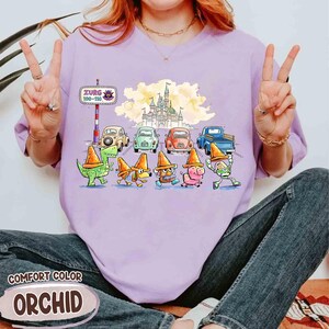 May include: Light purple Comfort Color t-shirt with a cartoon illustration. The design shows characters in traffic cones, vintage cars, and a castle. The text "Zurg 100-110" is on a sign. The shirt is a light purple colour.