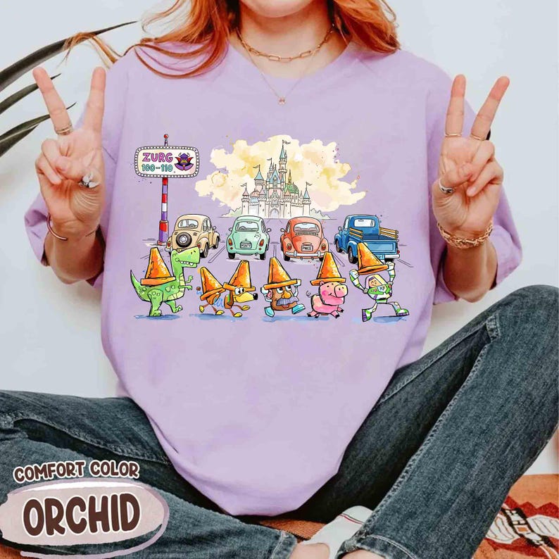 May include: Light purple Comfort Color t-shirt with a cartoon illustration. The design shows characters in traffic cones, vintage cars, and a castle. The text "Zurg 100-110" is on a sign. The shirt is a light purple colour.