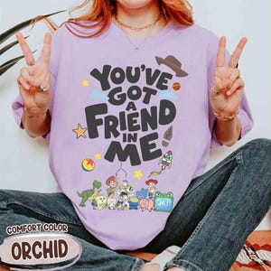 K&ouml;nnte beinhalten: Ein helllila T-Shirt mit dem Schriftzug "You've Got a Friend in Me" in einer verspielten Schriftart, mit Zeichentrickfiguren und einem Cowboyhut. Auf dem Shirt steht auch das Wort "Orchid".