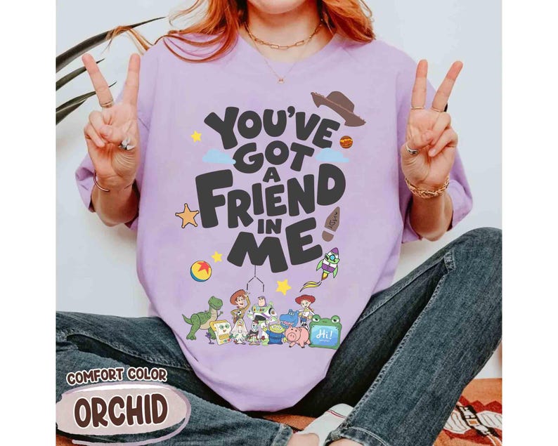 K&ouml;nnte beinhalten: Ein helllila T-Shirt mit dem Schriftzug "You've Got a Friend in Me" in einer verspielten Schriftart, mit Zeichentrickfiguren und einem Cowboyhut. Auf dem Shirt steht auch das Wort "Orchid".