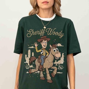 May include: Dark green t-shirt featuring a graphic of Sheriff Woody riding a horse, with the text "Sheriff Woody" in a cursive font. The design includes desert scenery and a waving Woody. The shirt has a white collar.