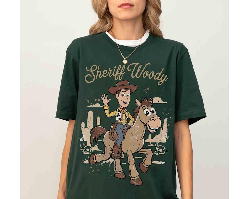 May include: Dark green t-shirt featuring a graphic of Sheriff Woody riding a horse, with the text "Sheriff Woody" in a cursive font. The design includes desert scenery and a waving Woody. The shirt has a white collar.