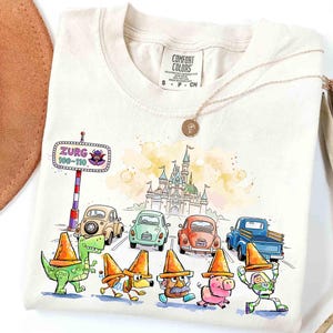 May include: Off-white t-shirt with a watercolour illustration of Disney characters and cars. The design includes a castle, a road sign that says "Zurg 100-110", and characters wearing orange traffic cones. A gold necklace and a brown hat are also visible.