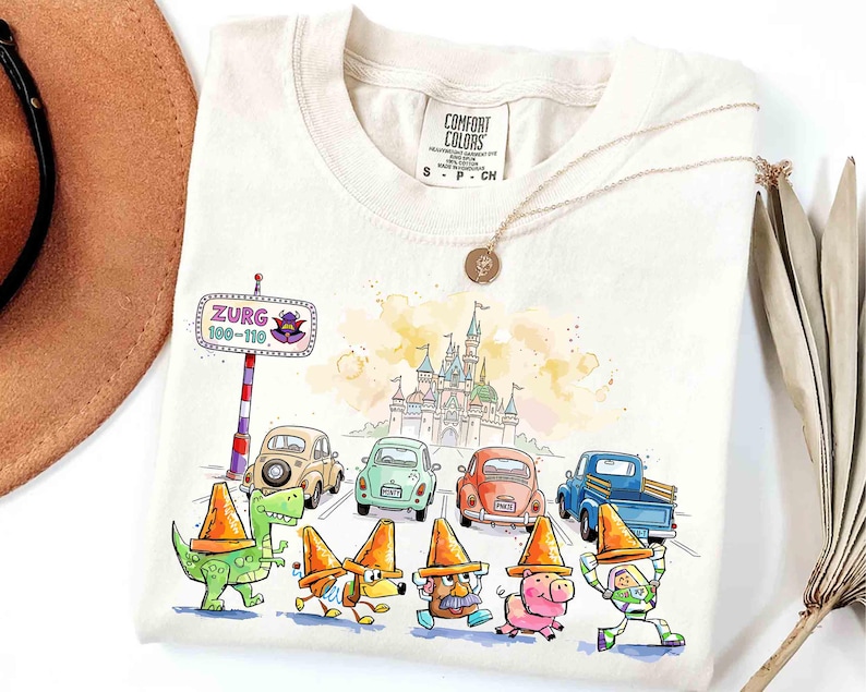May include: Off-white t-shirt with a watercolour illustration of Disney characters and cars. The design includes a castle, a road sign that says "Zurg 100-110", and characters wearing orange traffic cones. A gold necklace and a brown hat are also visible.