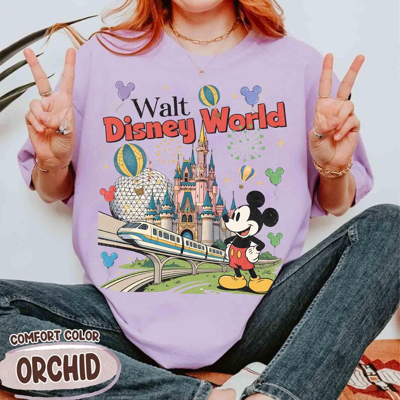 Puede incluir: Una camiseta lila Comfort Color con un gr&aacute;fico colorido de Walt Disney World. El dise&ntilde;o incluye un castillo, un tren, Mickey Mouse y globos. El texto "Walt Disney World" est&aacute; en rojo y amarillo.