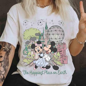 May include: Cream-colored t-shirt featuring Mickey and Minnie Mouse in front of a colorful cartoon landscape with a castle, sphere, and fireworks. The text "The Happiest Place on Earth" is printed below the image.