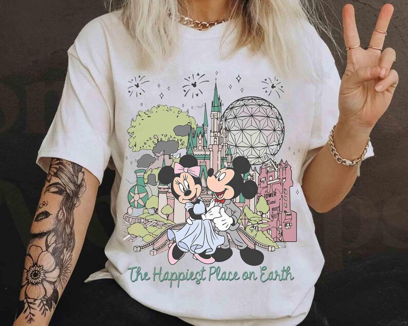 May include: Cream-colored t-shirt featuring Mickey and Minnie Mouse in front of a colorful cartoon landscape with a castle, sphere, and fireworks. The text "The Happiest Place on Earth" is printed below the image.