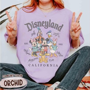 Puede incluir: Una camiseta lavanda con el logotipo de Disneyland y personajes de dibujos animados como Mickey Mouse, Minnie Mouse y Donald Duck. La camiseta incluye el texto "CALIFORNIA" y "EST. 1955". La camiseta est&aacute; etiquetada como "COMFORT COLOR ORCHID".