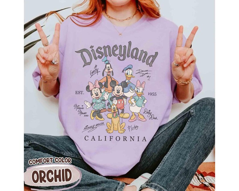 Puede incluir: Una camiseta lavanda con el logotipo de Disneyland y personajes de dibujos animados como Mickey Mouse, Minnie Mouse y Donald Duck. La camiseta incluye el texto "CALIFORNIA" y "EST. 1955". La camiseta est&aacute; etiquetada como "COMFORT COLOR ORCHID".