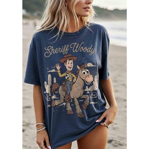 May include: A navy blue t-shirt featuring a graphic of Sheriff Woody riding a horse, with the text "Sheriff Woody" in a vintage font. The design includes desert scenery. The shirt is a casual, oversized style.