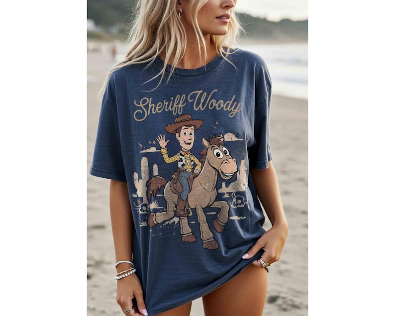 May include: A navy blue t-shirt featuring a graphic of Sheriff Woody riding a horse, with the text "Sheriff Woody" in a vintage font. The design includes desert scenery. The shirt is a casual, oversized style.