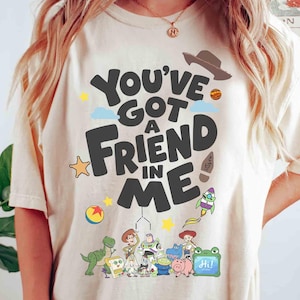 K&ouml;nnte beinhalten: Cremefarbenes T-Shirt mit dem Schriftzug "You've Got a Friend in Me" in einer verspielten Schriftart. Das Design umfasst Zeichentrickfiguren und Illustrationen eines Cowboyhuts, einer Rakete und eines Sterns. Ein farbenfrohes Grafik-T-Shirt.