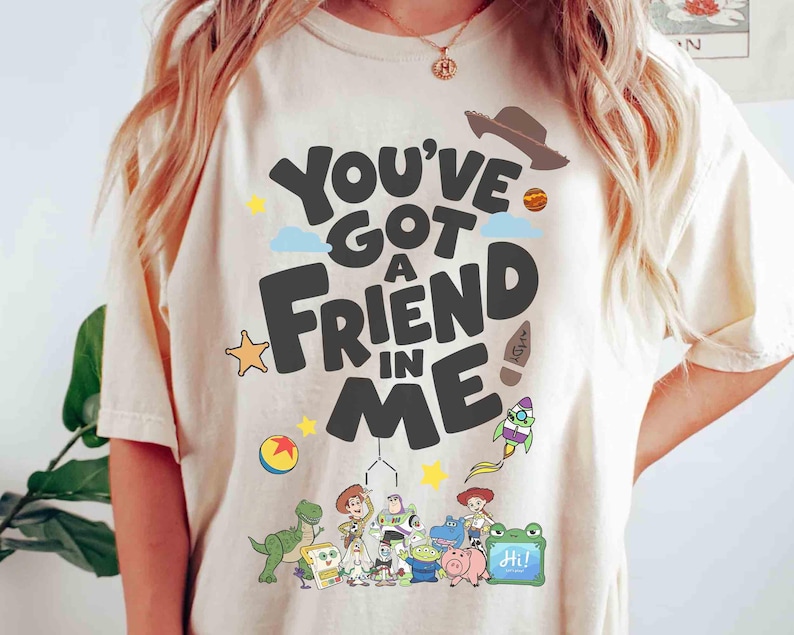 K&ouml;nnte beinhalten: Cremefarbenes T-Shirt mit dem Schriftzug "You've Got a Friend in Me" in einer verspielten Schriftart. Das Design umfasst Zeichentrickfiguren und Illustrationen eines Cowboyhuts, einer Rakete und eines Sterns. Ein farbenfrohes Grafik-T-Shirt.