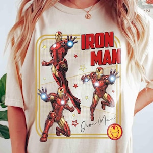 May include: A cream-coloured t-shirt with a graphic of Iron Man in action poses, and the words "Iron Man" in red block letters. The design includes yellow stars and a signature, all within a yellow rectangular border. The Iron Man logo is at the bottom.
