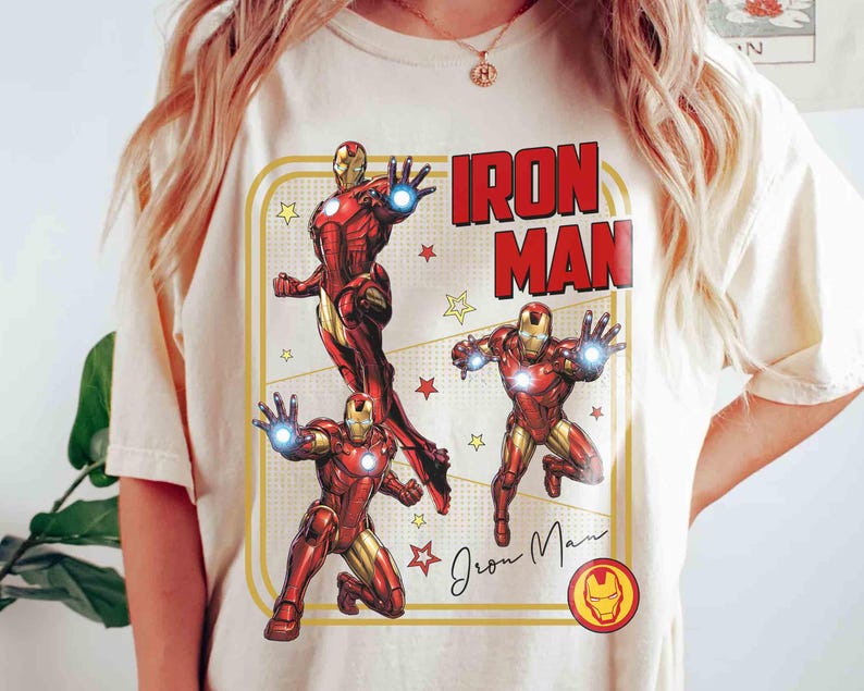 May include: A cream-coloured t-shirt with a graphic of Iron Man in action poses, and the words "Iron Man" in red block letters. The design includes yellow stars and a signature, all within a yellow rectangular border. The Iron Man logo is at the bottom.