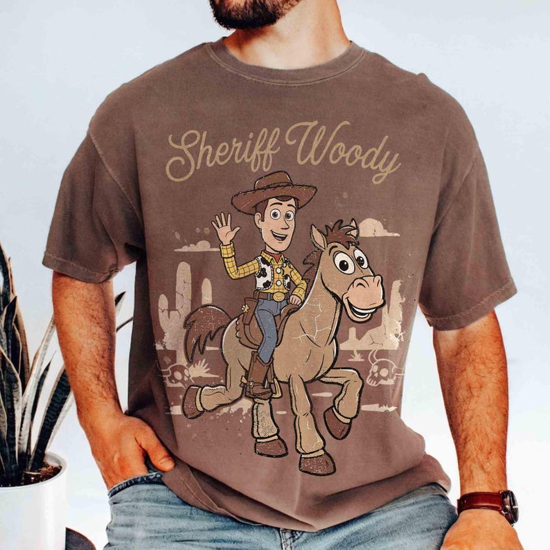 May include: Brown t-shirt featuring a cartoon illustration of Sheriff Woody riding a horse, with the text "Sheriff Woody" above the image. The design includes desert scenery and a waving Woody.