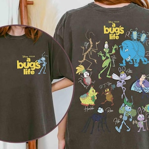 May include: Dark grey t-shirt featuring the Disney Pixar film "A Bug's Life." The front shows the movie title and an ant cartoon. The back displays various insect characters from the film in vibrant colours, including a blue rhinoceros beetle and a green grasshopper.