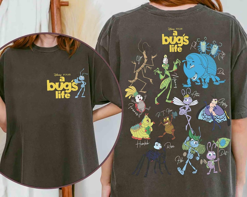 May include: Dark grey t-shirt featuring the Disney Pixar film "A Bug's Life." The front shows the movie title and an ant cartoon. The back displays various insect characters from the film in vibrant colours, including a blue rhinoceros beetle and a green grasshopper.