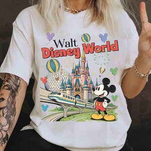 Puede incluir: Camiseta blanca con un gr&aacute;fico colorido de Walt Disney World. El dise&ntilde;o incluye el logotipo del parque, un castillo, Mickey Mouse, un monorra&iacute;l y globos. La camiseta tiene mangas cortas y cuello redondo.