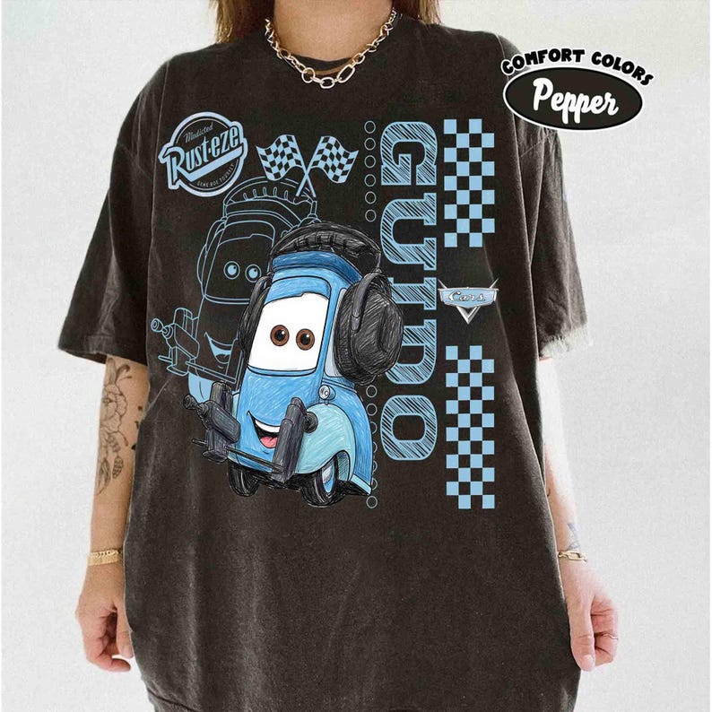 May include: Black Comfort Colors t-shirt featuring a cartoon image of Guido from the Cars movie. The design includes the text "Guido" in light blue, along with checkered flags and the "Rusteze" logo. The shirt has a distressed look.