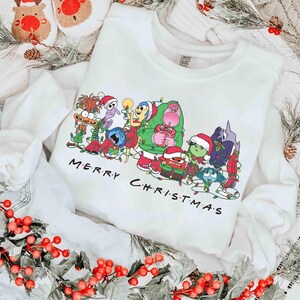 May include: White sweatshirt featuring a colorful graphic of animated characters in Christmas attire. The phrase "MERRY CHRISTMAS" is printed below the characters in black. The sweatshirt is surrounded by holiday decorations.