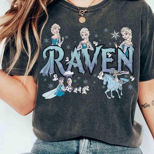 Puede incluir: Camiseta gris oscuro con la palabra "RAVEN" en letras azules y heladas, e ilustraciones de una princesa y copos de nieve. El dise&ntilde;o incluye una princesa montada en un caballo azul y otros personajes.