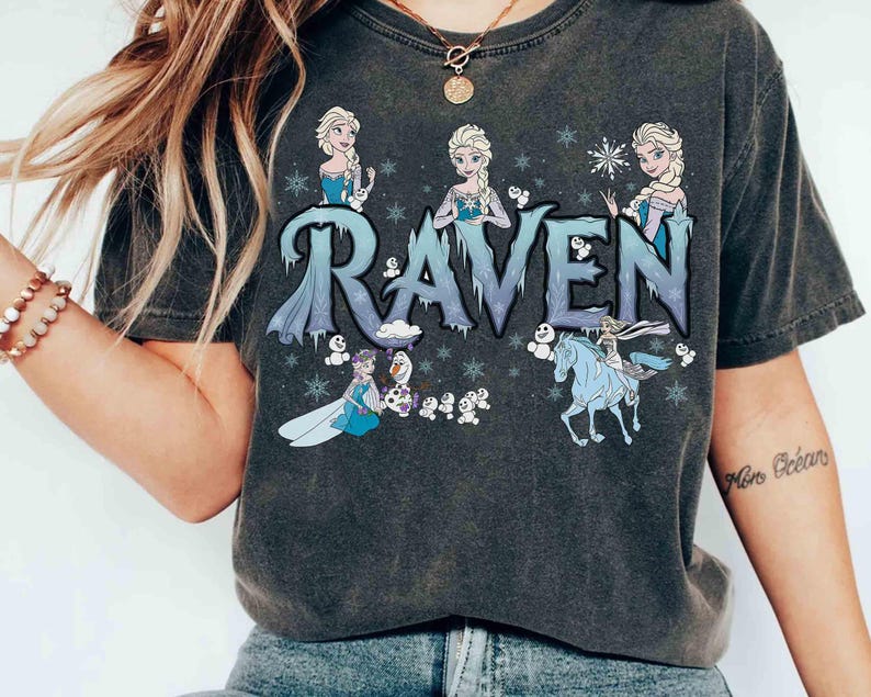Puede incluir: Camiseta gris oscuro con la palabra "RAVEN" en letras azules y heladas, e ilustraciones de una princesa y copos de nieve. El dise&ntilde;o incluye una princesa montada en un caballo azul y otros personajes.