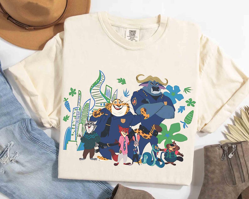 May include: Cream-coloured t-shirt with a vibrant graphic of characters from the animated film "Zootropolis." The design showcases animal characters in police uniforms and civilian clothes, set against a cityscape. The shirt has short sleeves and a crew neck.