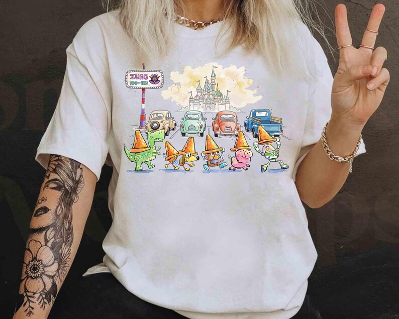 May include: White t-shirt with a cartoon design. The graphic shows a castle, cars, and characters wearing orange traffic cones. A sign reads "Zurg 100-110".