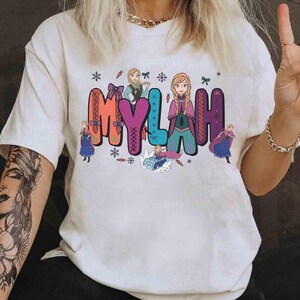 Puede incluir: Camiseta blanca con el nombre "MYLAH" en letras coloridas y estilizadas, acompa&ntilde;ada de personajes animados. El dise&ntilde;o incluye copos de nieve y personajes de dibujos animados, creando una est&eacute;tica l&uacute;dica y caprichosa.