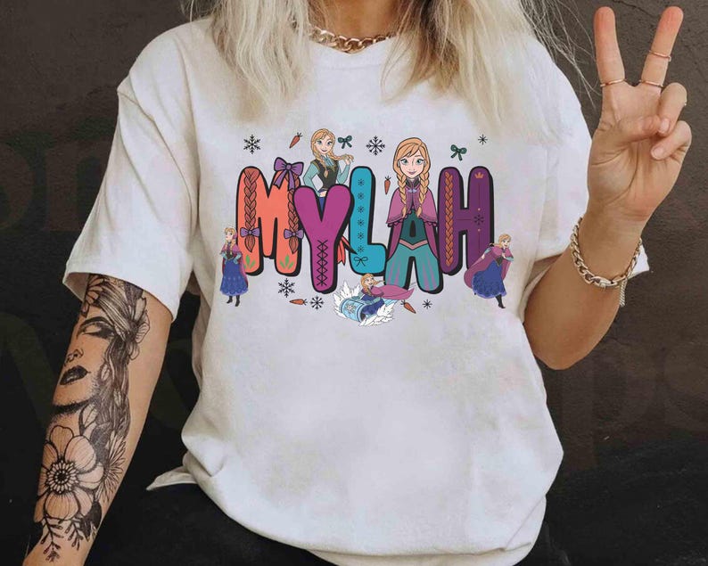 Puede incluir: Camiseta blanca con el nombre "MYLAH" en letras coloridas y estilizadas, acompa&ntilde;ada de personajes animados. El dise&ntilde;o incluye copos de nieve y personajes de dibujos animados, creando una est&eacute;tica l&uacute;dica y caprichosa.