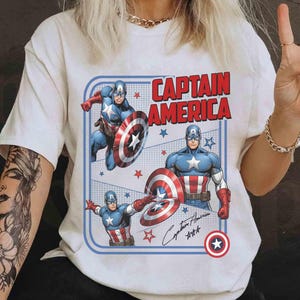 May include: White t-shirt with a Captain America design. The graphic shows Captain America, his shield, and the words "CAPTAIN AMERICA" in red. The design is framed with a blue border. The shirt has short sleeves.