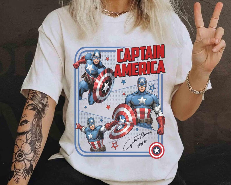 May include: White t-shirt with a Captain America design. The graphic shows Captain America, his shield, and the words "CAPTAIN AMERICA" in red. The design is framed with a blue border. The shirt has short sleeves.