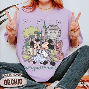 May include: Orchid-colored t-shirt featuring a graphic of Mickey and Minnie Mouse embracing in front of a castle and a geodesic sphere. The text "The Happiest Place on Earth" is below the graphic. The shirt is labeled "Comfort Color".