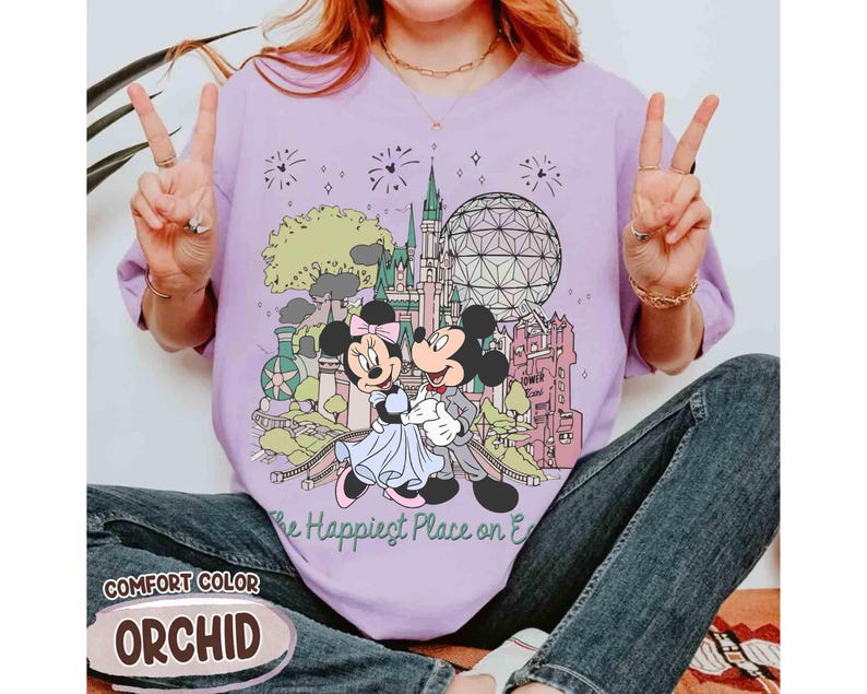 May include: Orchid-colored t-shirt featuring a graphic of Mickey and Minnie Mouse embracing in front of a castle and a geodesic sphere. The text "The Happiest Place on Earth" is below the graphic. The shirt is labeled "Comfort Color".
