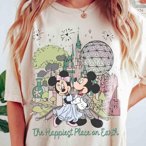 May include: Cream-colored t-shirt featuring a colorful graphic of Mickey and Minnie Mouse in front of a castle and a geodesic sphere. The text "The Happiest Place on Earth" is printed below the graphic. The shirt has short sleeves.
