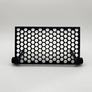 May include: A black rectangular stand featuring a repeating honeycomb pattern across its front surface. The structure is composed of a grid of hexagonal cutouts framed by a solid border, supported by two small, perpendicular base components.