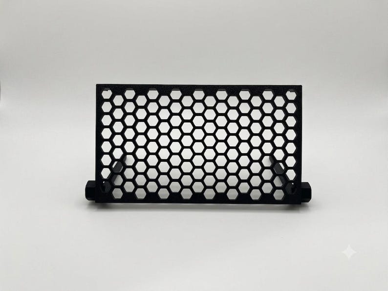 May include: A black rectangular stand featuring a repeating honeycomb pattern across its front surface. The structure is composed of a grid of hexagonal cutouts framed by a solid border, supported by two small, perpendicular base components.