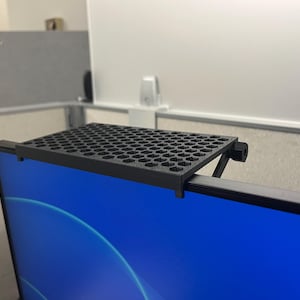 May include: A black monitor top shelf featuring a flat rectangular surface with a repeating hexagonal grid pattern. The shelf is designed to mount securely onto the upper edge of a computer screen, providing a stable platform for small items.
