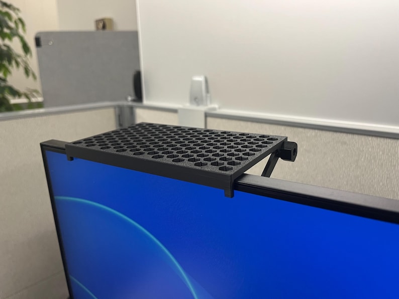 May include: A black monitor top shelf featuring a flat rectangular surface with a repeating hexagonal grid pattern. The shelf is designed to mount securely onto the upper edge of a computer screen, providing a stable platform for small items.