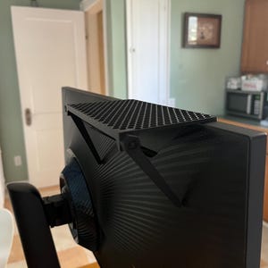 Puede incluir: Un estante superior para monitor de color negro con una superficie plana y rectangular con un patrón de panal perforado. El estante se fija al borde superior de un monitor de computadora con un brazo de soporte ajustable en la parte trasera.