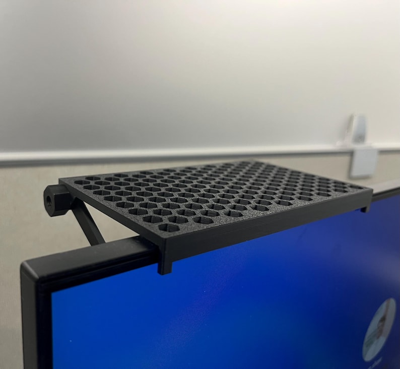 May include: A black monitor top shelf featuring a textured surface with a repeating hexagonal cutout pattern. The shelf is designed to mount onto the upper edge of a flat screen display, supported by an adjustable arm mechanism on the back.