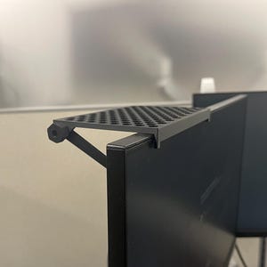 May include: A black monitor top shelf with a perforated surface and a support arm featuring a hexagonal adjustment knob. The shelf is mounted onto the top edge of a flat screen computer monitor, providing a flat horizontal surface for storage.