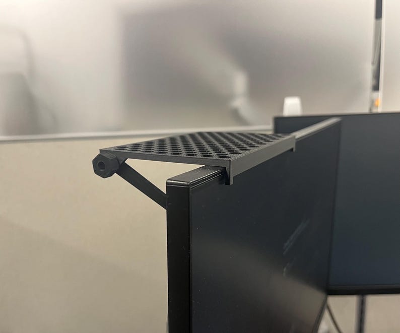 May include: A black monitor top shelf with a perforated surface and a support arm featuring a hexagonal adjustment knob. The shelf is mounted onto the top edge of a flat screen computer monitor, providing a flat horizontal surface for storage.