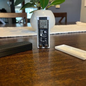 Puede incluir: Una placa de circuito electrónico con un diseño en blanco y negro, de pie. También hay una caja rectangular negra y una cubierta rectangular blanca. Los componentes están sobre una mesa de madera.