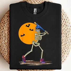 Puede incluir: Sudadera negra con un bateador esqueleto con casco y bate azules, sobre una luna naranja con siluetas de murciélagos. El esqueleto lleva zapatillas moradas y rojas. Diseño temático de Halloween.